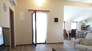 Un salon comprenant un canapé, une table basse et un téléviseur. Une porte-fenêtre coulissante donne accès à une cuisine avec un coin repas.