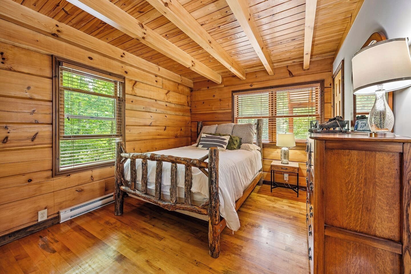 Un dormitorio de cabaña de madera con una cama rústica, dos ventanas con persianas, una mesita de noche y una lámpara.