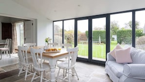 A dining area with a wooden table and white chairs, a sofa with cushions, and a view of a garden through large windows. - House Annexe Sleeps 18 Heated Pool (Chichester)