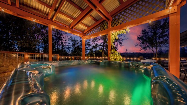 Un bain à remous sous une pergola en bois, avec vue sur les arbres et un coucher de soleil.