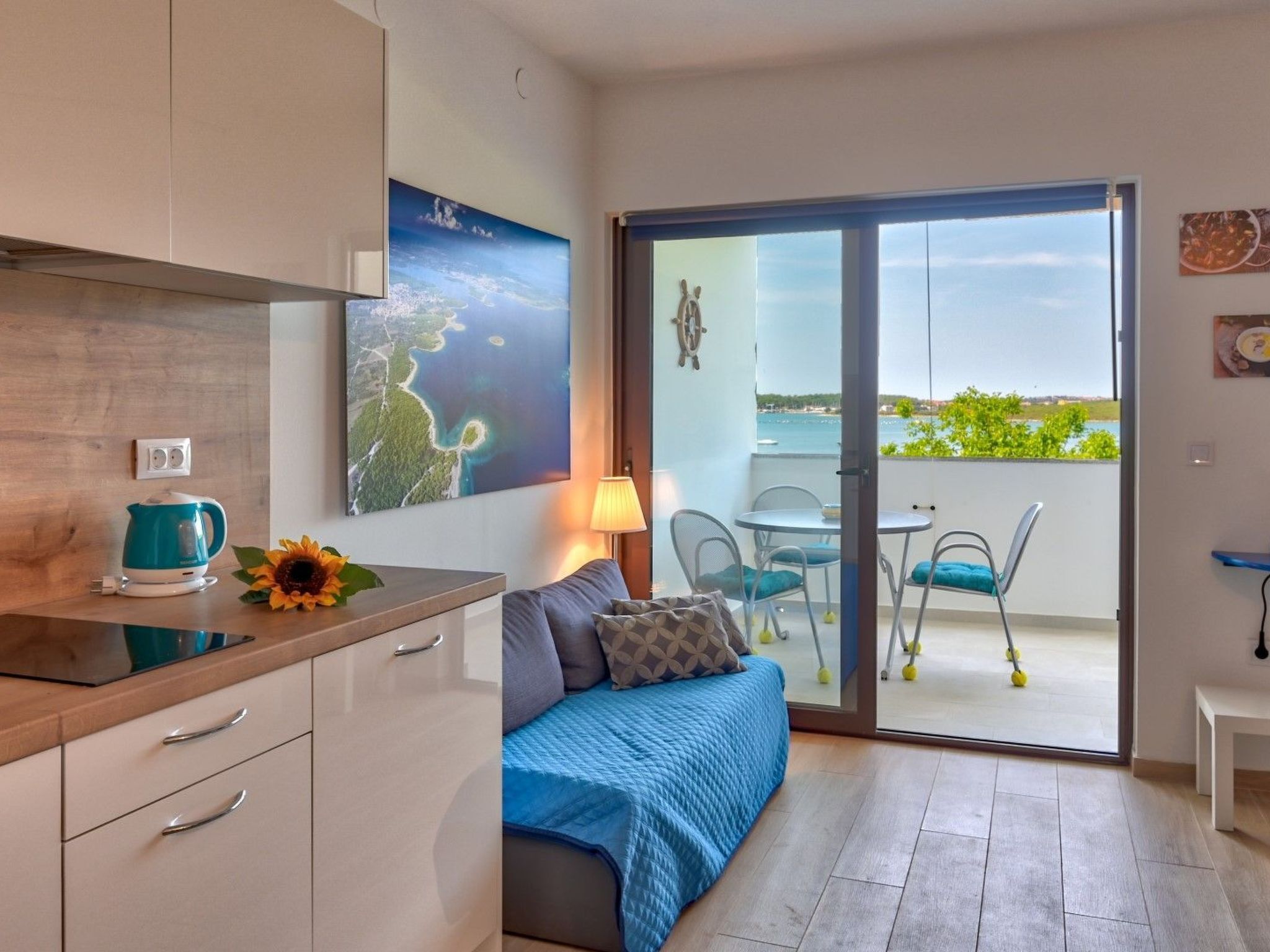 Une chambre moderne dotée d’une kitchenette, d’un canapé recouvert d’un plaid bleu, d’un coin repas avec une table et des chaises, et d’un balcon offrant une vue sur la mer.