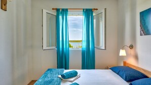Hotellihuone, jossa on sänky, kaksi tyynyä, ikkuna verhoilla, seinään kiinnitetty valaisin ja taulu.