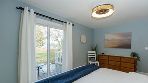 Ein Schlafzimmer mit einem Bett, einer Holzkommode, einem Sessel, einem Bild an der Wand und Blick ins Freie durch eine Schiebetür.