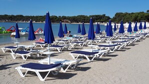 Ein Strand mit Reihen aus weißen Liegestühlen und blauen Sonnenschirmen, einem klaren Himmel und einem ruhigen Meer.