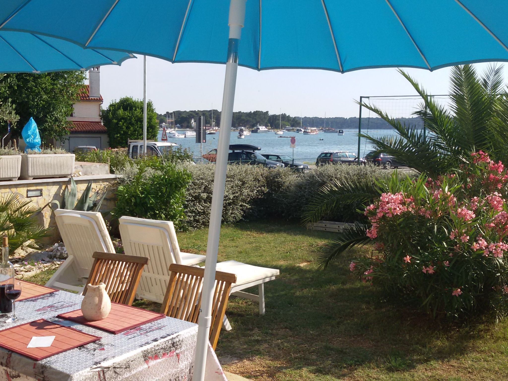 Zona de estar al aire libre con una mesa y sillas bajo un paraguas azul, con vista a un puerto deportivo con barcos.