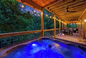 Uma banheira de hidromassagem numa varanda de madeira, com vista para árvores, um ventilador de teto e luzes de corda.