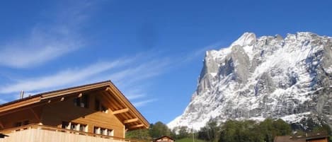Edificio de estilo chalé con fachada de madera y un balcón, frente a una montaña nevada y un cielo azul despejado.