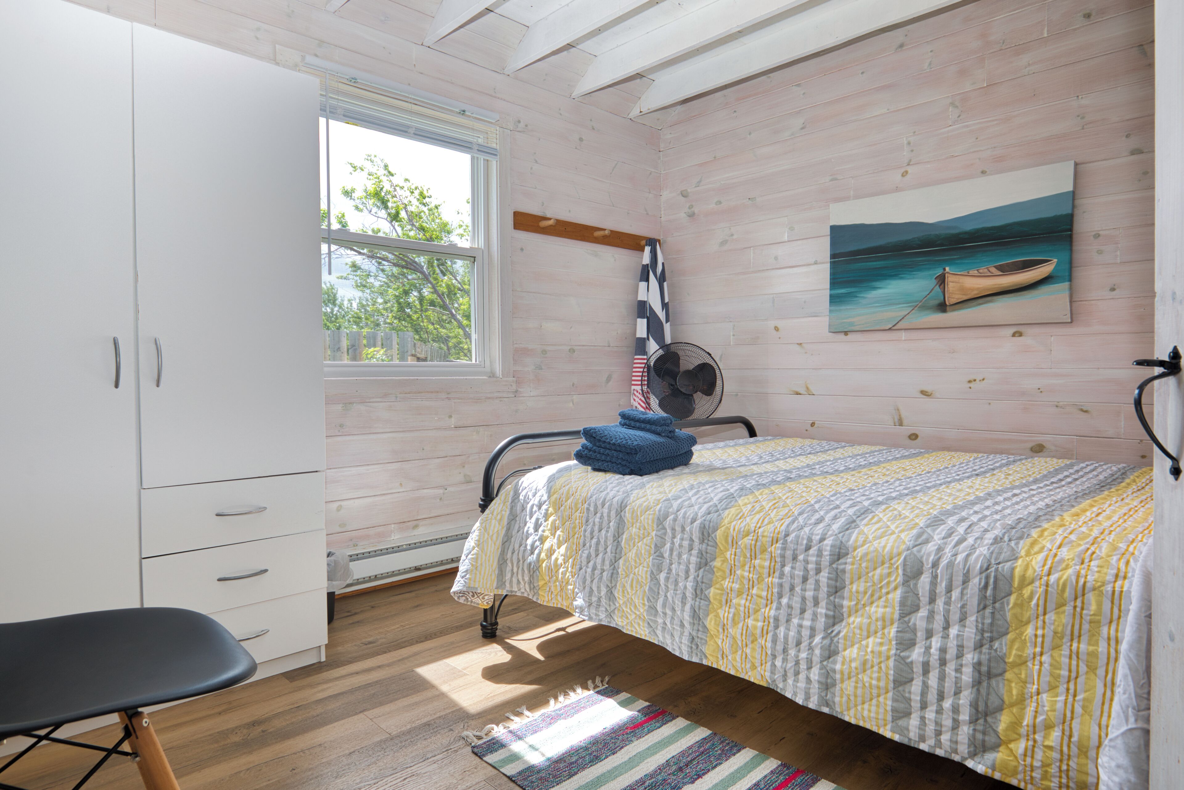 Un dormitorio con una cama, un escritorio, una silla, un ventanal, un ventilador y un cuadro en la pared.