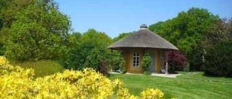 A small, single-story building with a thatched roof, surrounded by lush greenery and yellow flowering plants.