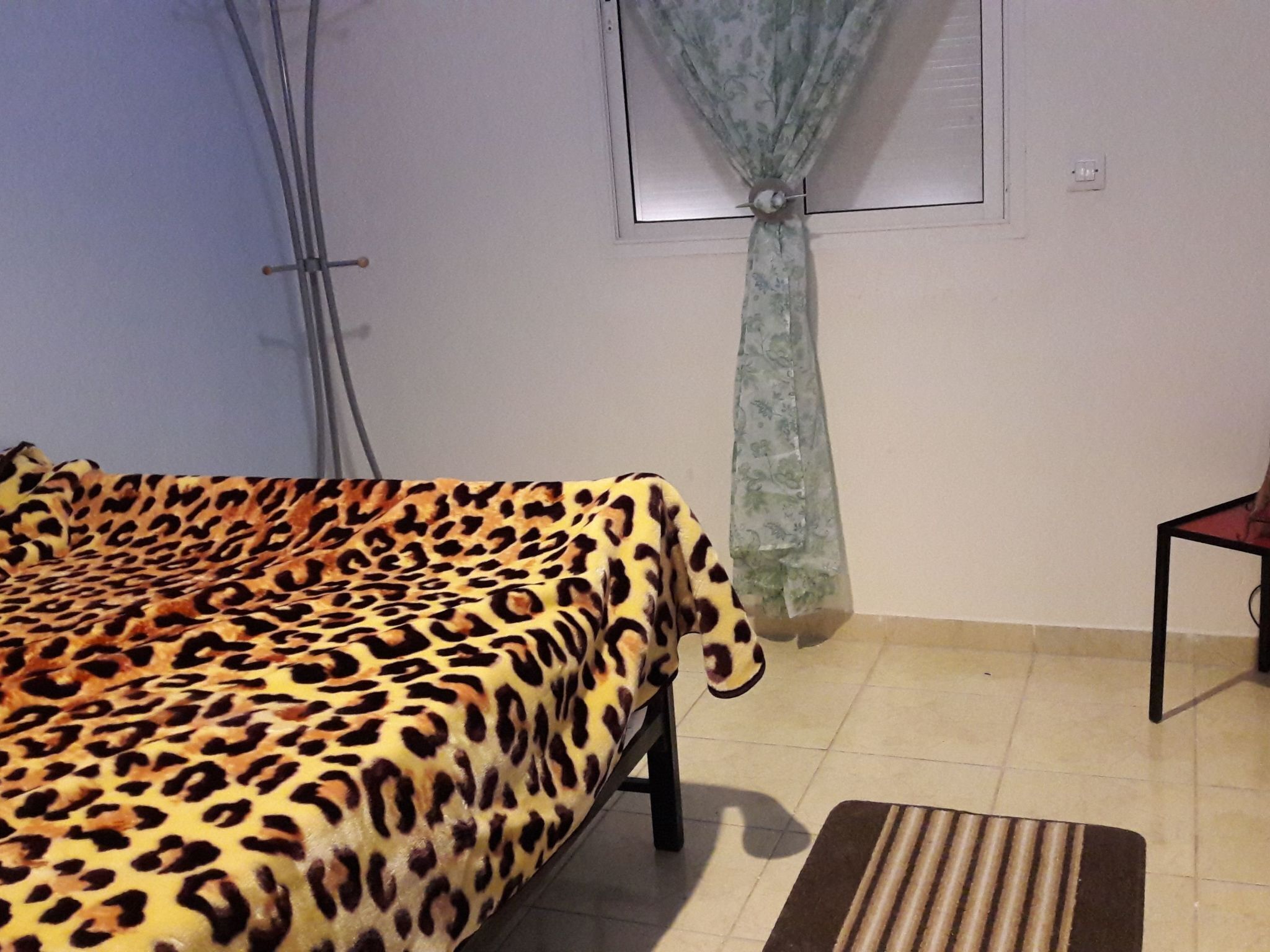 A room with a bed covered in a leopard print blanket, a window with a curtain, and a small table with a chair.