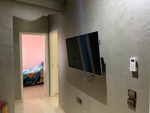 Un couloir avec un téléviseur fixé au mur, une porte donnant sur une pièce où se trouve un lit, et un thermostat également fixé au mur.