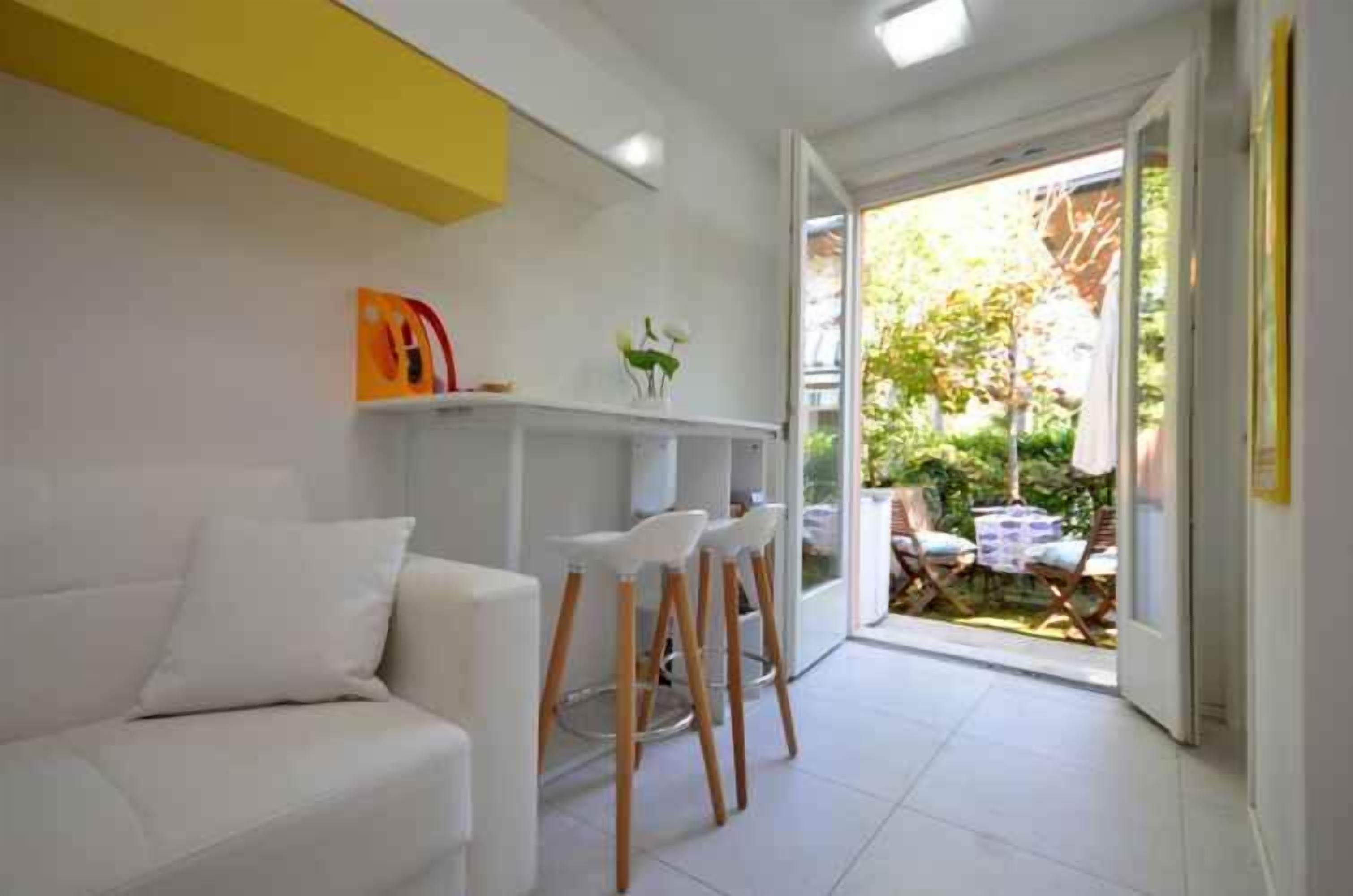 A modern living room with a white sofa, a small bar area with stools, and a sliding glass door leading to an outdoor seating area.
