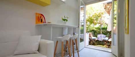 A modern living room with a white sofa, a small bar area with stools, and a sliding glass door leading to an outdoor seating area.