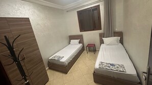 Ein kleines, sauberes Zimmer mit zwei Einzelbetten, einem kleinen Tisch, einem Fenster mit Vorhängen und einem Kleiderständer.