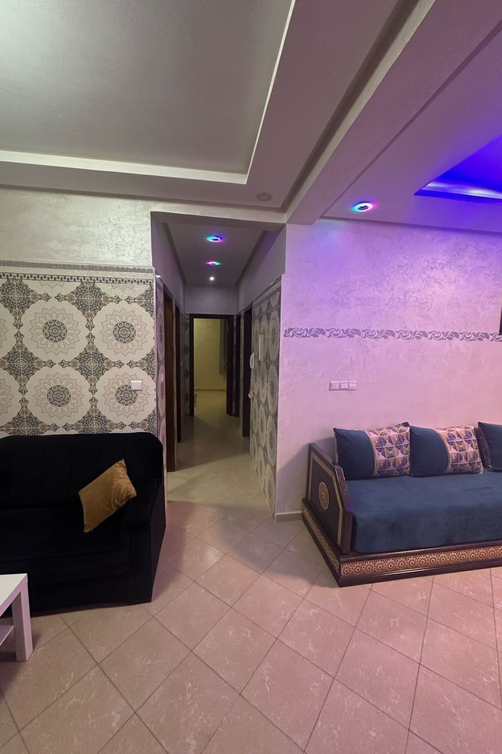 A living room with a black sofa and a blue sofa with patterned cushions, a patterned wall, and a hallway with a door leading to another room.