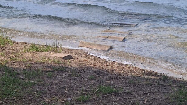 Ein Strand mit klarem Wasser, einem sandigen Ufer und einer fernen Küstenlinie mit Bäumen und Gebäuden.