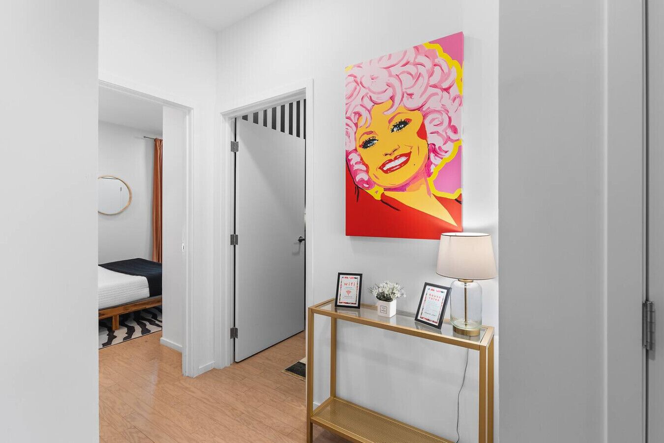Un couloir avec un lit, une porte, un tableau encadré et une lampe posée sur une console.