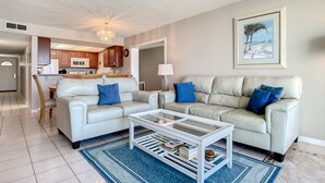 A modern living room with a white leather sofa, a glass coffee table, and a blue rug. There is a kitchen in the background with wooden cabinets and a microwave. - Oceanfront Retreat on No-Drive Beach | Clean, Peaceful & Perfectly Located (New Smyrna Beach)