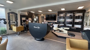 Interior de salón con mostrador de recepción que dice "Sandra Zysko", un espejo, sillas y estantes con productos para el cabello.