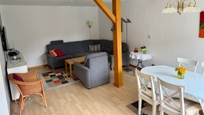 Een moderne woonkamer met een grijze bank, een plafond met houten lijstwerk, een eettafel met witte stoelen en een bijzettafeltje met een lamp.