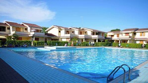 A swimming pool surrounded by apartment buildings with tiled walkways and a clear blue sky. - Holiday apartment with air conditioning and balcony (Bibione)