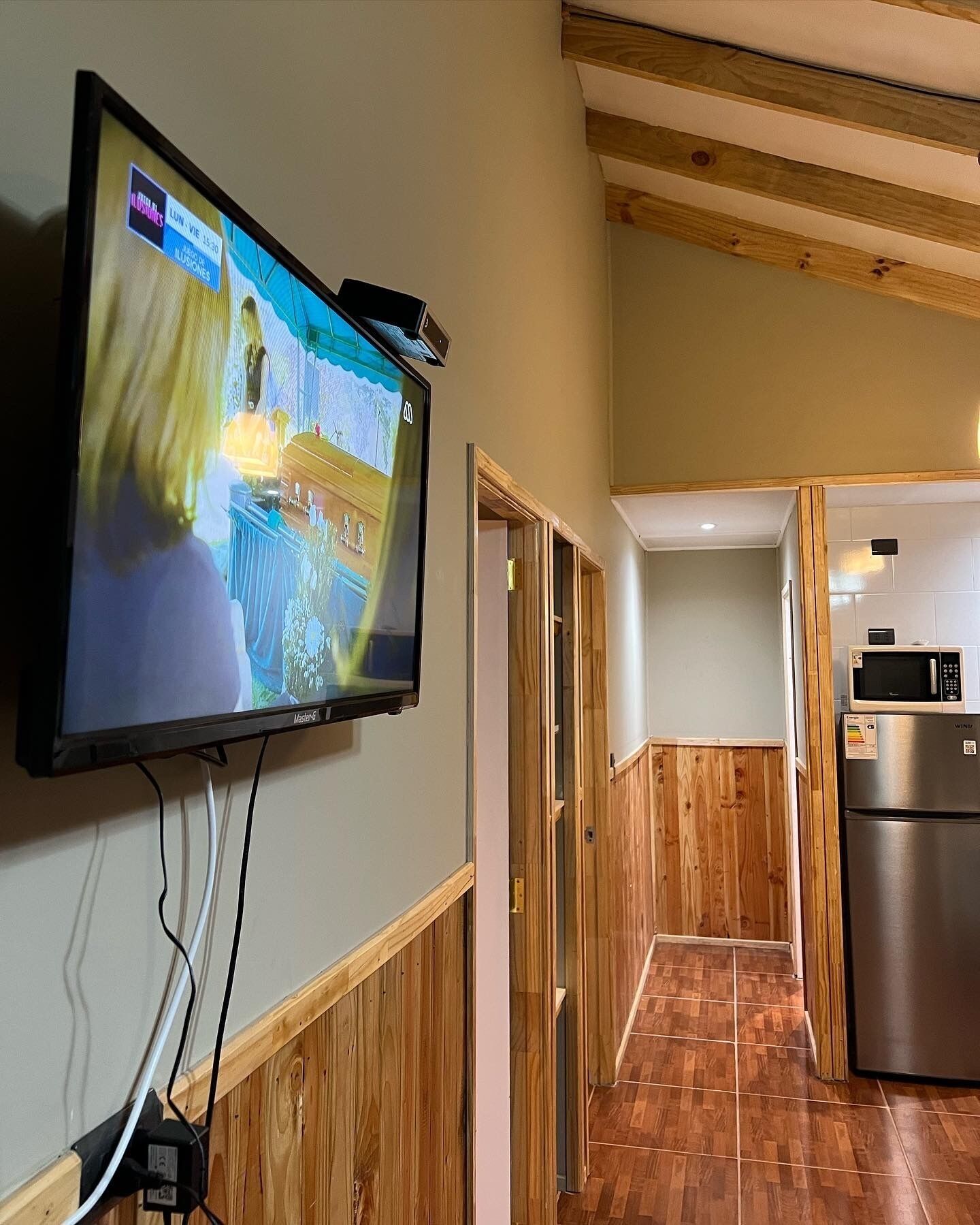 A modern kitchen with a stainless steel refrigerator, wooden flooring, and a flat-screen TV mounted on the wall.