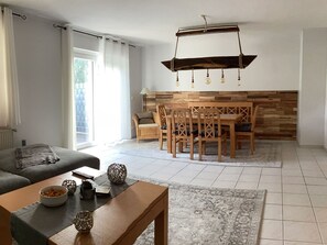 Een moderne woonkamer met een bank, een houten eettafel met stoelen, een kroonluchter en een tapijt met een patroon.