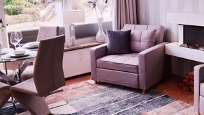 A modern living room with a grey sofa, a dining table with chairs, a patterned rug, and a view of greenery outside. - Strandappartment by Interhome (Glücksburg)