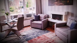 A modern living room with a fireplace, a dining area with a glass table, and a large window offering a view of the outdoors. - Strandappartment by Interhome (Glücksburg)
