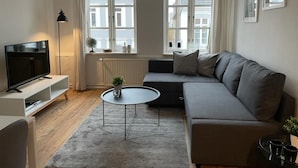 Un soggiorno moderno con un divano grigio, una TV a schermo piatto su un mobile bianco, un tavolino rotondo e due finestre con tende bianche.