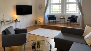 Ein modernes Wohnzimmer mit Flachbildfernseher, zwei Sofas, einem Glastisch und einem großen Fenster mit Vorhängen.