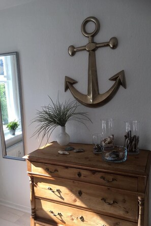 Een houten dressoir met laden, een decoratief anker aan de muur, een potplant en een glazen vaas met schelpen.