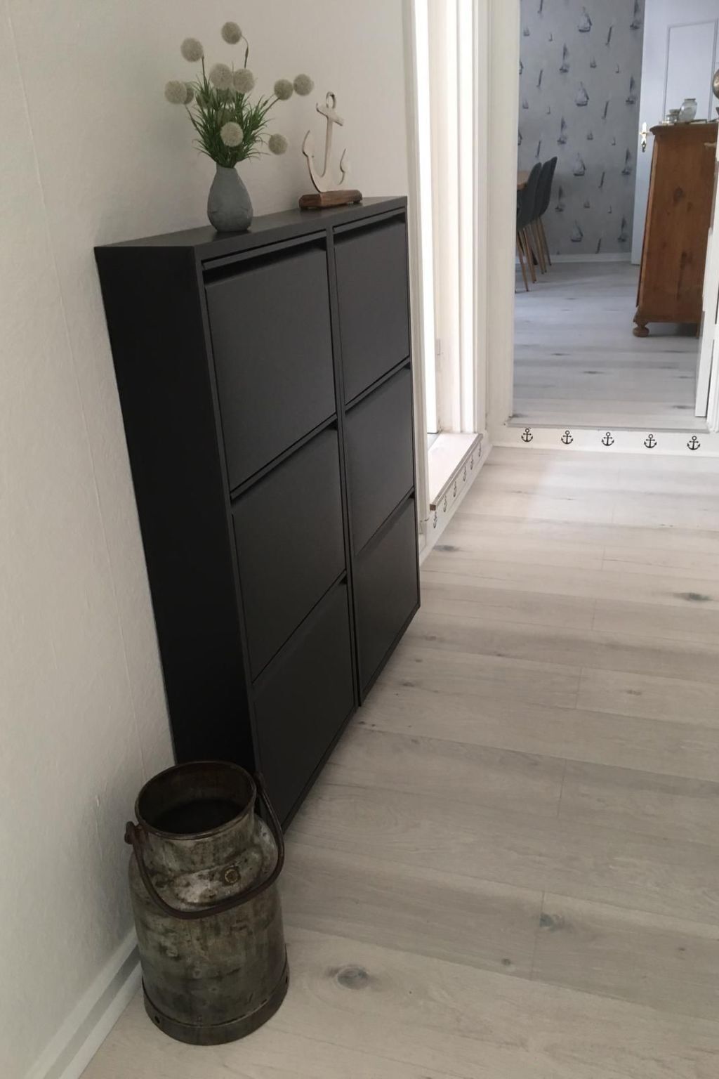 A room with a dark wooden cabinet against a white wall, a rustic metal bucket, and a patterned wallpaper in the background.