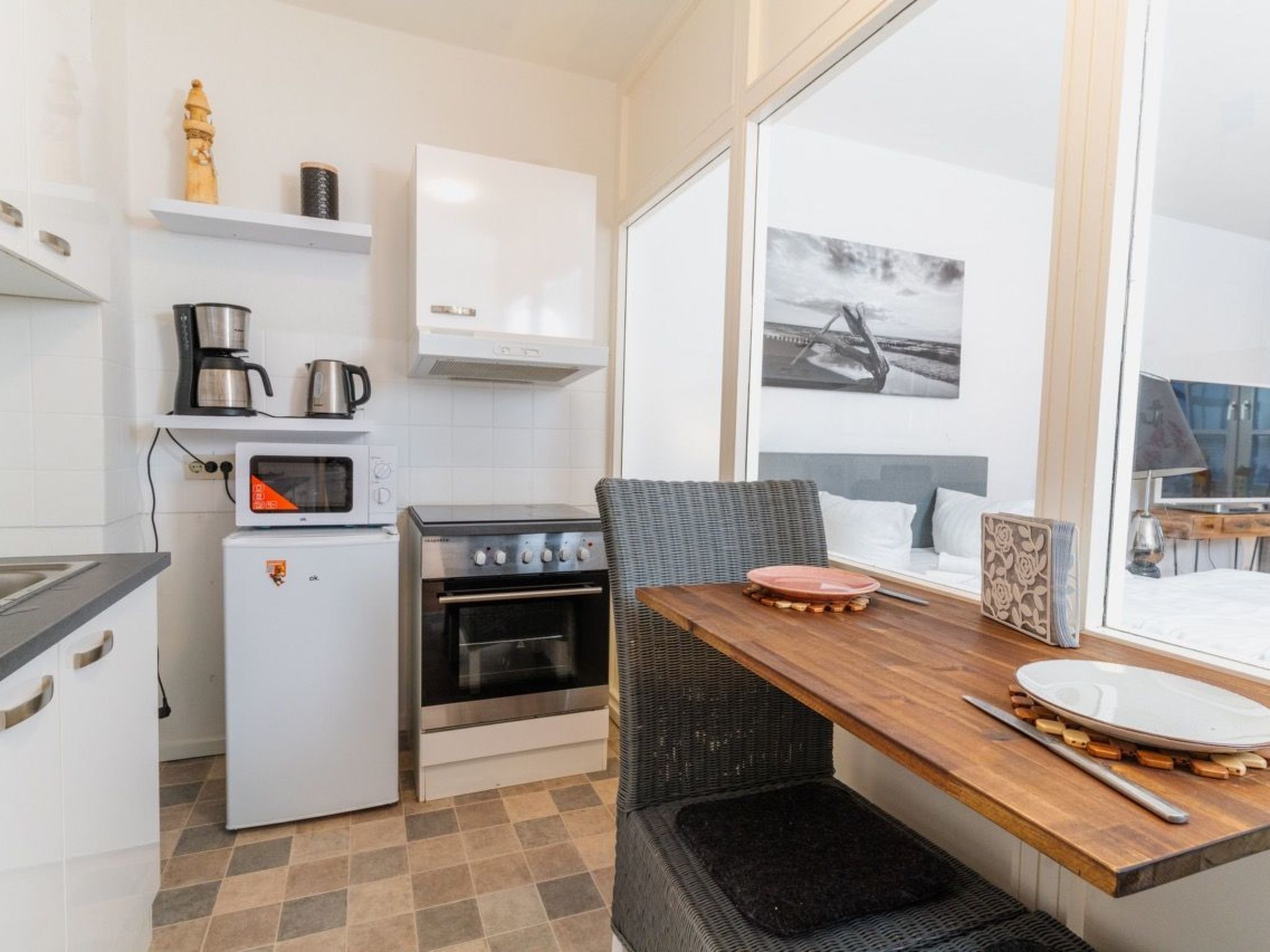 A compact kitchen with a microwave, coffee maker, and stove, a dining area with a wooden table and wicker chair, and a large window with a view of a bridge.
