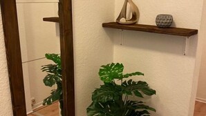 Um canto com espelho de madeira, planta monstera em vaso e prateleira de madeira com objetos decorativos.
