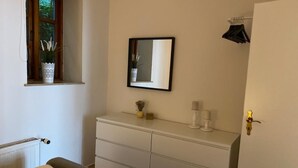 Quarto com cama, cómoda, espelho, janela com plantas e radiador.