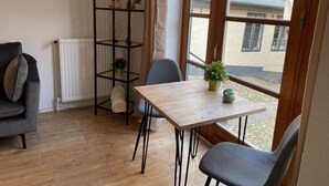 Quarto com piso de madeira, prateleira de metal preto com objetos decorativos, mesa com cadeiras e uma janela grande com vista para edifícios.