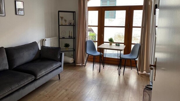 Sala de estar com um sofá cinza escuro, uma mesinha com duas cadeiras, uma estante de livros e uma janela com cortinas.