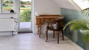 Quarto com escrivaninha de madeira, cadeira, radiador e janela grande com vista para áreas verdes.