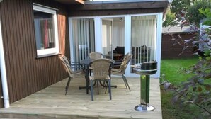 Una terraza de madera con una mesa y sillas, un pequeño jardín y un abrevadero metálico para pájaros.