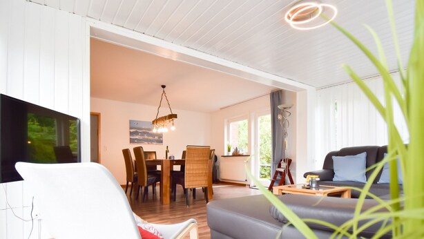 Una sala moderna con un sofá gris, una silla blanca, una mesa de comedor con sillas y un televisor montado en la pared.