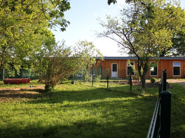 A wooden building with a door and windows, surrounded by a fenced area with grass and trees.