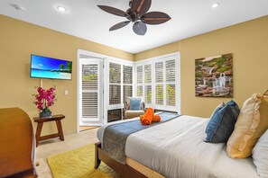 Ein Schlafzimmer mit einem Bett, einem Fernseher, einem Deckenventilator, einem Fenster mit Jalousien und einem Bild an der Wand.