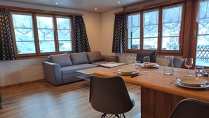 Una sala de estar moderna con un sofá, una mesa de comedor y sillas. Hay ventanas amplias con cortinas, y vistas a un paisaje nevado.