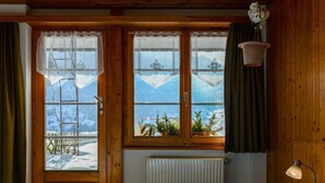 Una habitación con paredes de madera, una cama con colcha de cuadros azules y blancos, un radiador y una ventana con vistas a montañas y un paisaje nevado.