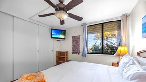 A hotel room with a bed, a ceiling fan, a TV, a window with curtains, and a small table with a lamp. - Napili Point | Oceanfront 2 BR, Sleeps 8 | Car Incl. w/ 6+ Nights | NAP ML-3494 by KBM (Lahaina)