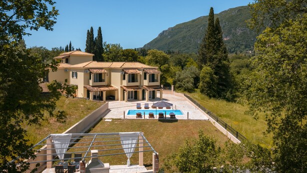 Uma casa de dois andares com piscina, rodeada por vegetação e colinas.