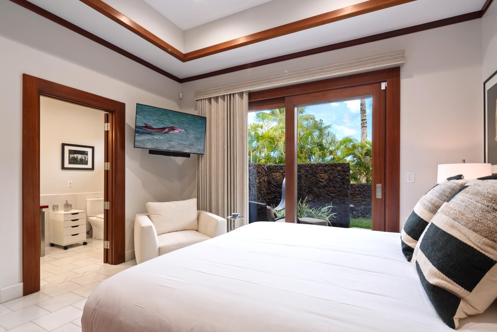 A bedroom with a large bed, a television, a chair, and a view of a tropical outdoor area.