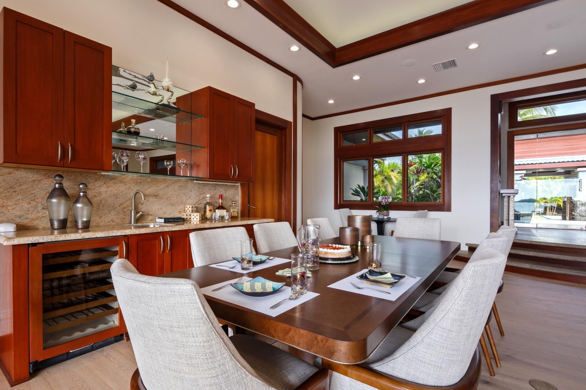 A spacious dining area with a large wooden table, white upholstered chairs, and a kitchen with wooden cabinets and a glass display shelf.
