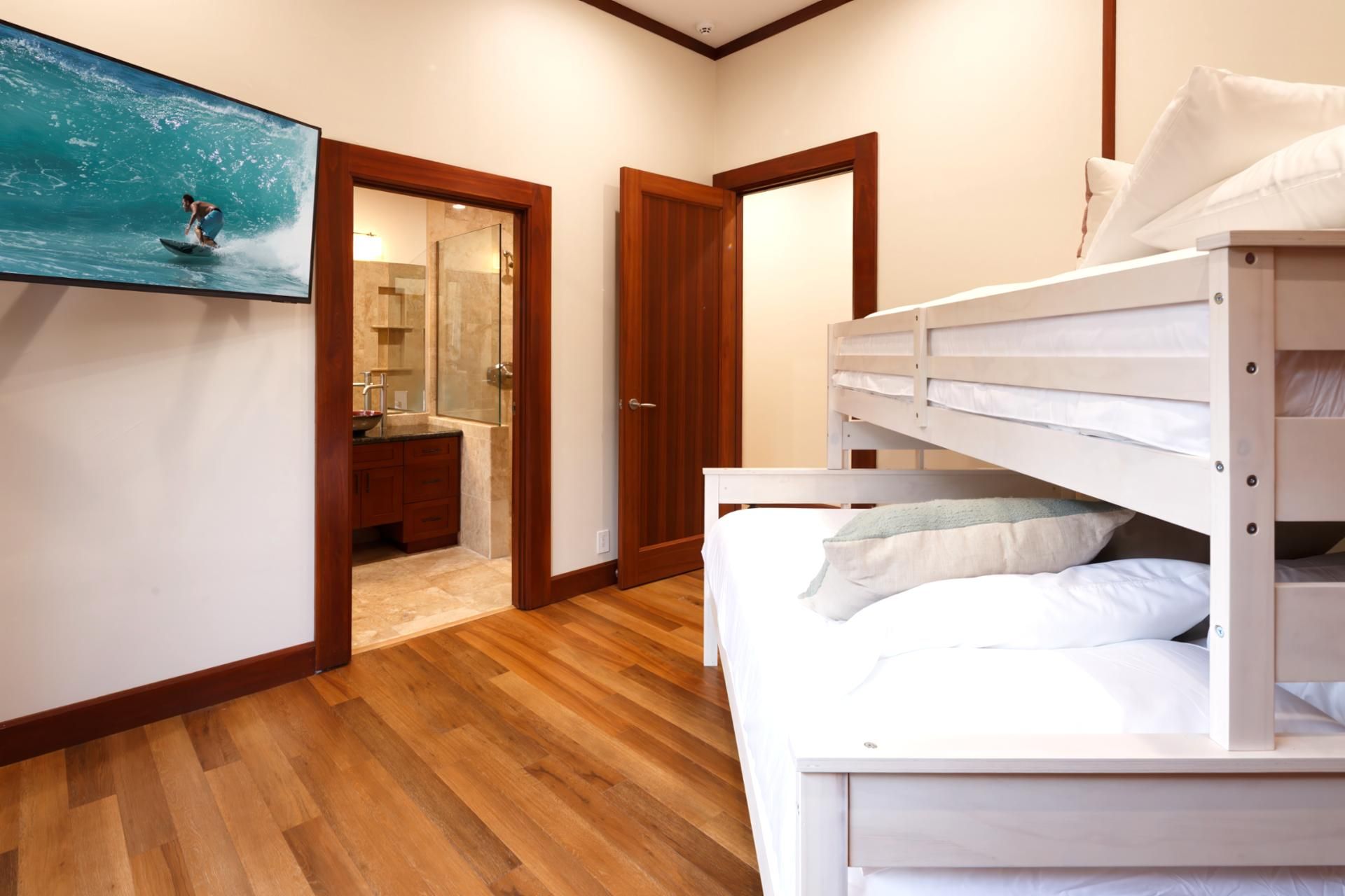 A room with a bunk bed, wooden flooring, a bathroom with a glass shower, and a painting of a surfer on a wave.